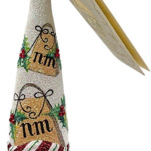May include: A Christmas ornament shaped like Santa Claus. The ornament is white with glitter and features a red and white striped hat, a white beard, and the letters "nm" on a gold background. The ornament is decorated with green leaves and red berries.