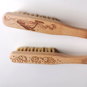 May include: Two wooden brushes with natural bristles. The first brush has a whale design burned into the wood. The second brush has a design of three turtles burned into the wood.