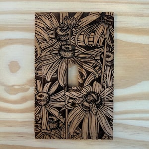 May include: Wooden light switch cover with a laser-etched floral design featuring large flowers and a bee. The cover is rectangular with a single switch opening. The wood has a natural, light brown color with dark brown etched details.