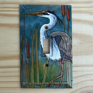 May include: A rectangular light switch plate featuring a painted illustration of a grey and white heron standing in a marsh with green reeds and brown cattails. The artwork covers the entire surface, including the central toggle switch cutout.