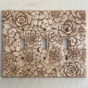 May include: A wooden light switch plate with a detailed pyrography design of succulent plants. The plate has four switch openings and is finished with a natural wood stain. The design features various sizes of succulents.