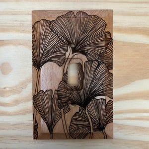 May include: A wooden light switch cover featuring a detailed, etched design of ginkgo leaves. The leaves are black and the cover is a light brown color. The switch opening is rectangular.