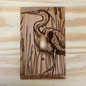 May include: Wooden light switch cover featuring an engraved heron standing in a marsh. The heron is detailed with etched feathers and a long beak. Cattails and reeds are also etched into the wood, creating a nature-themed design.