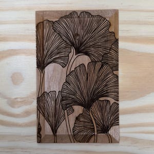 May include: A rectangular wooden plaque featuring a detailed, dark brown laser-etched design of ginkgo leaves. The leaves are arranged in a stylized pattern, with visible veins and a natural wood grain background. The plaque has a small hole at the top.