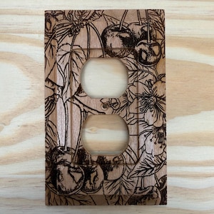 May include: Wooden double outlet cover with a cherry blossom design. The cover features two circular openings for outlets and is decorated with detailed, dark-brown etched cherries, leaves, and flowers. The wood has a natural, light-brown color.
