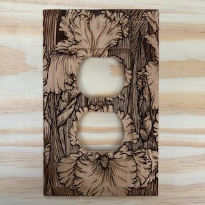 May include: Wooden double outlet cover with a detailed floral design. The cover features two circular openings for outlets and is decorated with etched iris flowers. The wood has a natural, light brown color with darker brown etched details.