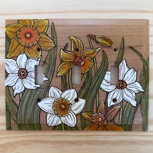 May include: A rectangular wooden triple light switch cover plate featuring a hand-painted illustration of yellow and white daffodils with green stems and leaves. The design includes three vertical rectangular cutouts for toggle switches.