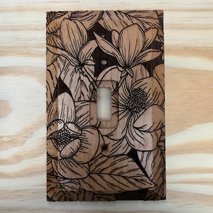 May include: Wooden light switch cover with a floral design. The cover features a detailed engraving of magnolia flowers and leaves. The light switch opening is rectangular. The wood has a natural, light brown color with darker brown engraved details.