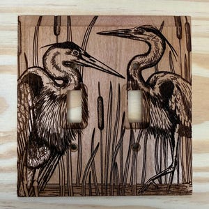 May include: Wooden double light switch cover featuring a detailed pyrography design of two herons standing in a marsh. The artwork is in shades of brown and black, with cattails and reeds in the background.