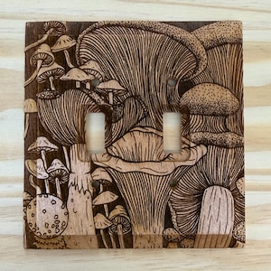 May include: Wooden double light switch cover featuring an intricate carved design of various mushrooms. The cover is made of light and dark brown wood, with the mushroom details highlighted by the wood grain.