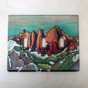 May include: A light switch cover with a watercolor painting of a mountain range. The mountains are painted in shades of red, orange, yellow, blue, and green. The switch cover has three toggle switches.
