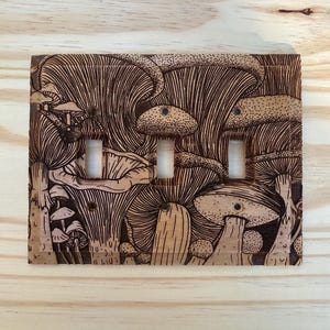 May include: A rectangular wooden light switch plate featuring a detailed pyrography design of various mushrooms. The artwork covers the entire surface, incorporating three vertical rectangular cutouts for switches and four small screw holes.