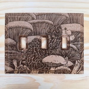 May include: A wooden light switch cover featuring a detailed engraved illustration of various mushrooms. The design includes different types of fungi, with intricate line work creating a textured appearance. The cover has three switch openings and is mounted on a light wood surface.