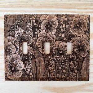 May include: Wooden light switch cover with a floral design. The cover features a detailed engraving of pansies and lily of the valley flowers. The cover has three switch openings and is made of light brown wood.