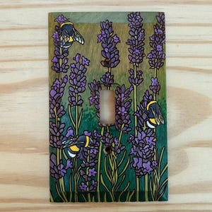 May include: A rectangular wooden light switch cover plate featuring a hand-painted design of purple lavender stalks and three yellow and black bumblebees. The background is a gradient of green and brown tones with a central rectangular cutout.