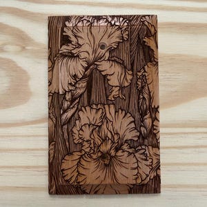 May include: A wooden light switch cover featuring a detailed, engraved design of iris flowers. The flowers are depicted in a naturalistic style with intricate lines and shading, creating a three-dimensional effect. The cover is rectangular with two holes for the switch.