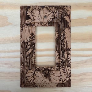 May include: A wooden light switch cover featuring a floral pattern. The rectangular cover has a single switch aperture. The floral design is etched into the wood, providing a detailed, textured appearance. The wood has a natural, light brown colour.