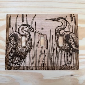 May include: Wooden light switch cover featuring a pyrography design of two herons standing in a marsh with cattails. The birds are detailed with dark brown shading, set against a light brown background.