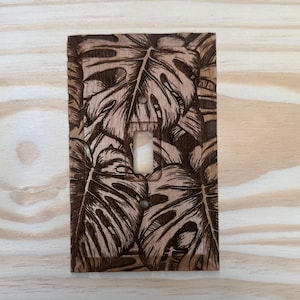 Peut inclure: Plaque d'interrupteur en bois avec un motif de pyrogravure de feuilles de monstera. Les feuilles sont gravées dans le bois, créant un thème botanique naturel. La plaque a une ouverture pour un interrupteur et deux trous de vis.