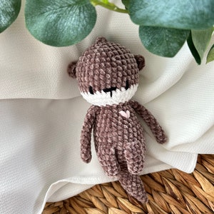 Könnte beinhalten: Ein braunes, gehäkeltes Plüschtier in Form eines Otters mit weißem Gesicht und einem rosa Herz auf der Brust. Das Spielzeug liegt auf einer weißen Stoffoberfläche mit einem geflochtenen Korb darunter.