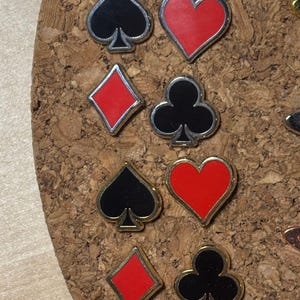 May include: Close-up of playing card suit pins. The pins feature the shapes of spades, hearts, diamonds, and clubs. The spades and clubs are black, while the hearts and diamonds are red. Each pin has a silver or gold border.