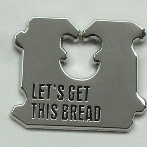 Puede incluir: Un cuchillo para pan de metal plateado con un mango con forma de pan. El mango tiene un recorte en forma de rebanada de pan. El cuchillo tiene el texto "Let's Get This Bread" grabado en el mango.