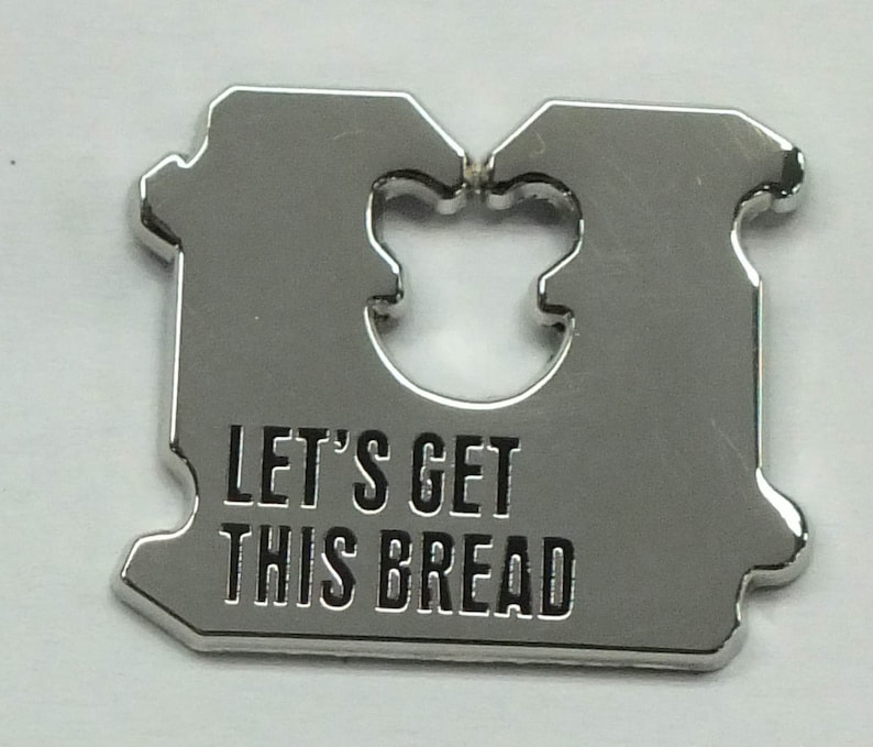 Puede incluir: Un cuchillo para pan de metal plateado con un mango con forma de pan. El mango tiene un recorte en forma de rebanada de pan. El cuchillo tiene el texto "Let's Get This Bread" grabado en el mango.