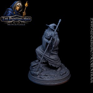 May include: A gray resin miniature of a robed figure wearing a pointed hat and holding a long staff. The figure stands on a rocky, circular base. The text THE PAINTING MAGE MINIATURES appears in the top left corner with a logo of a hooded figure.
