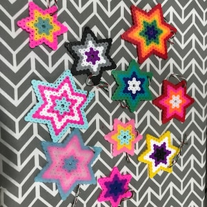 May include: A collection of colorful star-shaped ornaments made from small, fused plastic beads. The ornaments feature various color schemes, including rainbow, pink, blue, and yellow. Each star has a loop for hanging.