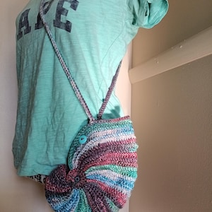 May include: A handmade, multi-colored crochet purse with a button closure and strap. The purse features a spiral design with shades of blue, green, purple, and pink. The strap is also crocheted and matches the purse's color scheme.