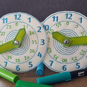 Könnte beinhalten: Zwei weiße, kreisförmige Lernuhren mit blauen Zahlen und grünen Zeigern. Die Uhren haben ein 24-Stunden-Format und sind mit blauen Stichen bestickt. Grüne und blaue Marker befinden sich im Vordergrund.