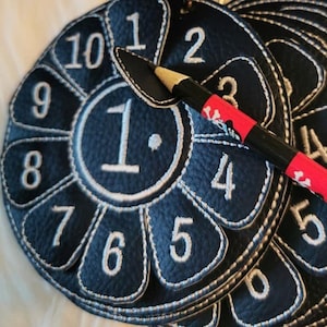 May include: A black leather game piece with white stitching and numbers 1 through 10. The piece is shaped like a flower with 10 petals. A pencil with a red and white striped design is lying on top of the game piece.