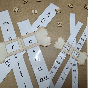 May include: Two wooden bone-shaped frames with cut-out windows for letter tiles. The frames are labeled "re" and "ma wo".  The frames are placed on top of strips of paper with letters printed on them, including "m l n", "s f t", "l u", "au", "si", "re le", "wa no fu", "ne ja jo", "ni je pi", "lu pa mo", "se wi so", "la mu fo", "ja nu pe".