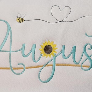 May include: Embroidered design of the word "August" in teal blue script with a sunflower, a bee, a heart, and cherries.