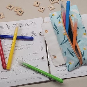 Könnte beinhalten: Eine blaue Stiftebox mit einem weißen Blumenmuster und einem Reißverschluss. Die Stiftebox ist geöffnet und enthält mehrere farbige Stifte und Bleistifte.