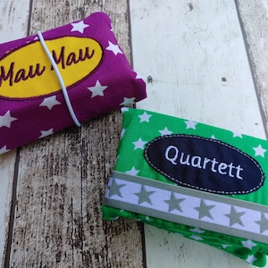 May include: Two fabric pouches with white stars. One pouch is purple with a yellow oval that says "Mau Mau". The other pouch is green with a black oval that says "Quartett".