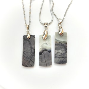 May include: Three rectangular stone pendants hanging from silver-toned metal chains. Each pendant features a unique natural pattern with shades of grey, white, and dark charcoal veining. The stones are polished and held by silver-toned bails.