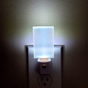 May include: A rectangular white night light plugged into a wall socket. The device features a flat, translucent, rectangular panel that emits a bright, cool-toned white glow, illuminating the surrounding area with a soft, diffused light.