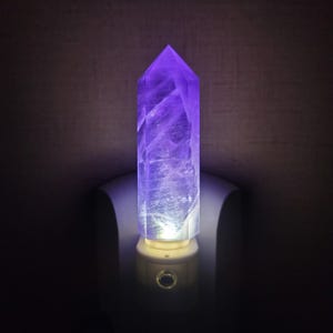 May include: A tall, purple crystal night light. The crystal is a light purple color and is lit from the base, creating a soft glow. The night light is plugged into a wall outlet.