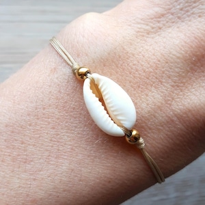 Op de afbeelding: Een delicate schelpenarmband. De armband heeft een natuurlijke witte schelp als middelpunt, geregen op een dun, beige koord. Kleine goudkleurige kralen accentueren de schelp. De armband is ontworpen om om de pols te worden gedragen.