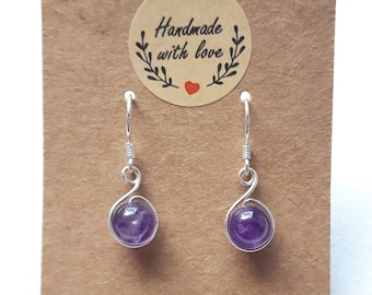 May include: A pair of silver dangle earrings with purple amethyst stones. The earrings are handmade and feature a delicate wire design. The card reads "Handmade with love".