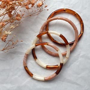 Könnte beinhalten: Vier Armbänder in Braun-, Rosa- und Cremetönen. Die Armbänder haben ein segmentiertes Design mit goldfarbenen Akzenten. Getrocknete Blumen befinden sich in der oberen linken Ecke.