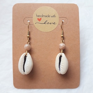 Peut inclure: Une paire de boucles d'oreilles dorées avec une perle brune et une coquille de cauris blanche suspendue. Les boucles d'oreilles sont présentées sur une carte brune avec un autocollant fait main avec amour.