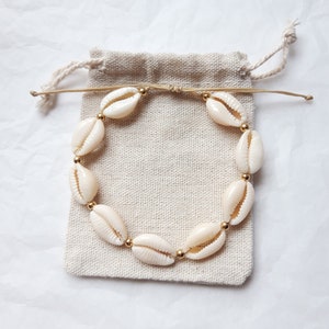 Peut inclure: Un bracelet en coquillages blancs avec des perles dorées sur un fond en tissu beige.