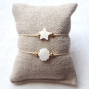 Peut inclure: Deux bracelets délicats avec des perles dorées et des breloques en coquillage blanc. Un charme est une étoile et l'autre est un coquillage. Les bracelets sont présentés sur une surface en tissu beige.