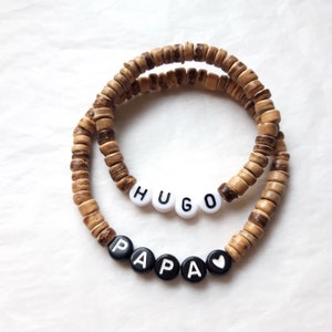 Può includere: Due braccialetti di perline di legno con i nomi "HUGO" e "PAPA" scritti in perline bianche e nere. Il braccialetto "PAPA" ha anche una piccola perlina a forma di cuore bianca.