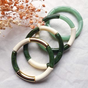 Op de afbeelding: Drie kleurrijke armbanden. Eén armband is groen en wit met gouden accenten. Een andere is groen en de derde is lichtgroen. De armbanden zijn gemaakt van afgeronde segmenten. Gedroogde bloemen staan op de achtergrond.