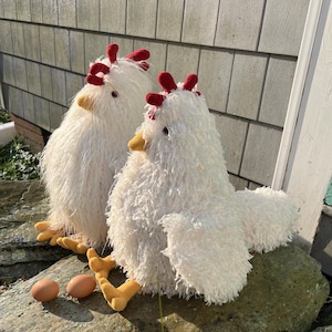 May include: Two white fluffy plush chickens with red combs and yellow beaks sitting on a rock with two brown eggs in front of them.