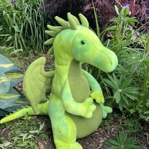 May include: A plush green dragon toy with a long neck, wings, and a tail. The dragon is in a seated position, with its arms resting on its belly. The dragon is surrounded by green plants and foliage.