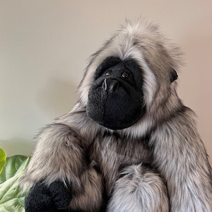 以下が含まれることがあります： ふわふわの毛並みのグレーとブラックのゴリラのぬいぐるみ。ゴリラは籐のバスケットに座っています。